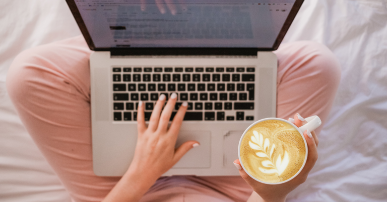 A person sits cross-legged with a laptop on their lap, planning an email series. They type with one hand while holding a latte with leaf-shaped foam art in the other, all against a light-colored bed or blanket backdrop.