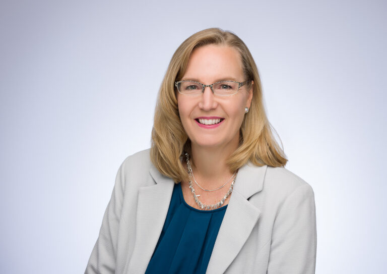A direct response copywriter with blonde hair, wearing glasses, a light gray blazer, teal blouse, and a silver necklace, smiles at the camera against a plain light background.