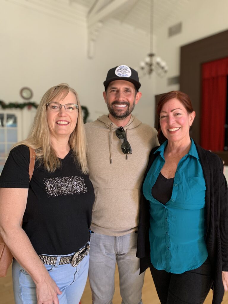 Three adults stand close together indoors, smiling at the camera—perfectly capturing a friendly networking moment. The woman on the left wears glasses and a black shirt, while the others wear a cap with beige hoodie and a teal blouse with black cardigan.