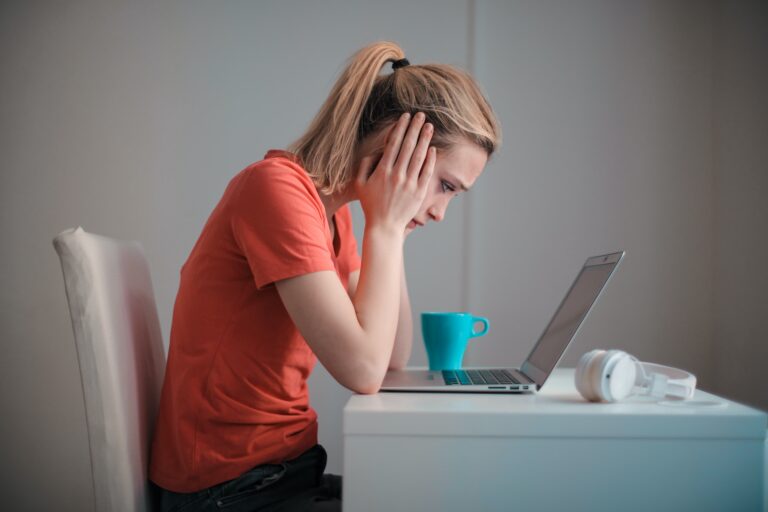 A woman with a ponytail sits at a desk, looking stressed while staring at her laptop. She rests her head in her hands, struggling with indecision over her online marketing strategy. A blue mug and white headphones are on the table beside her.