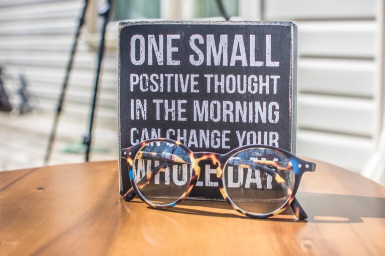 A pair of tortoiseshell eyeglasses rests on a wooden table in front of a sign that reads, One small positive thought in the morning can help you keep your focus while serving clients at the highest level.