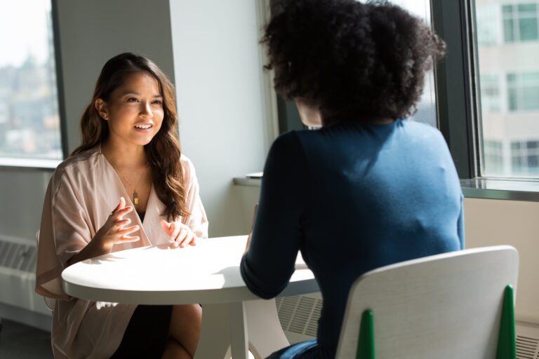 Two women sit across from each other at a round table by a window, engaged in conversation. One woman faces the camera, sharing an online lead generation tip and gesturing with her hands, while the other has her back to the camera.