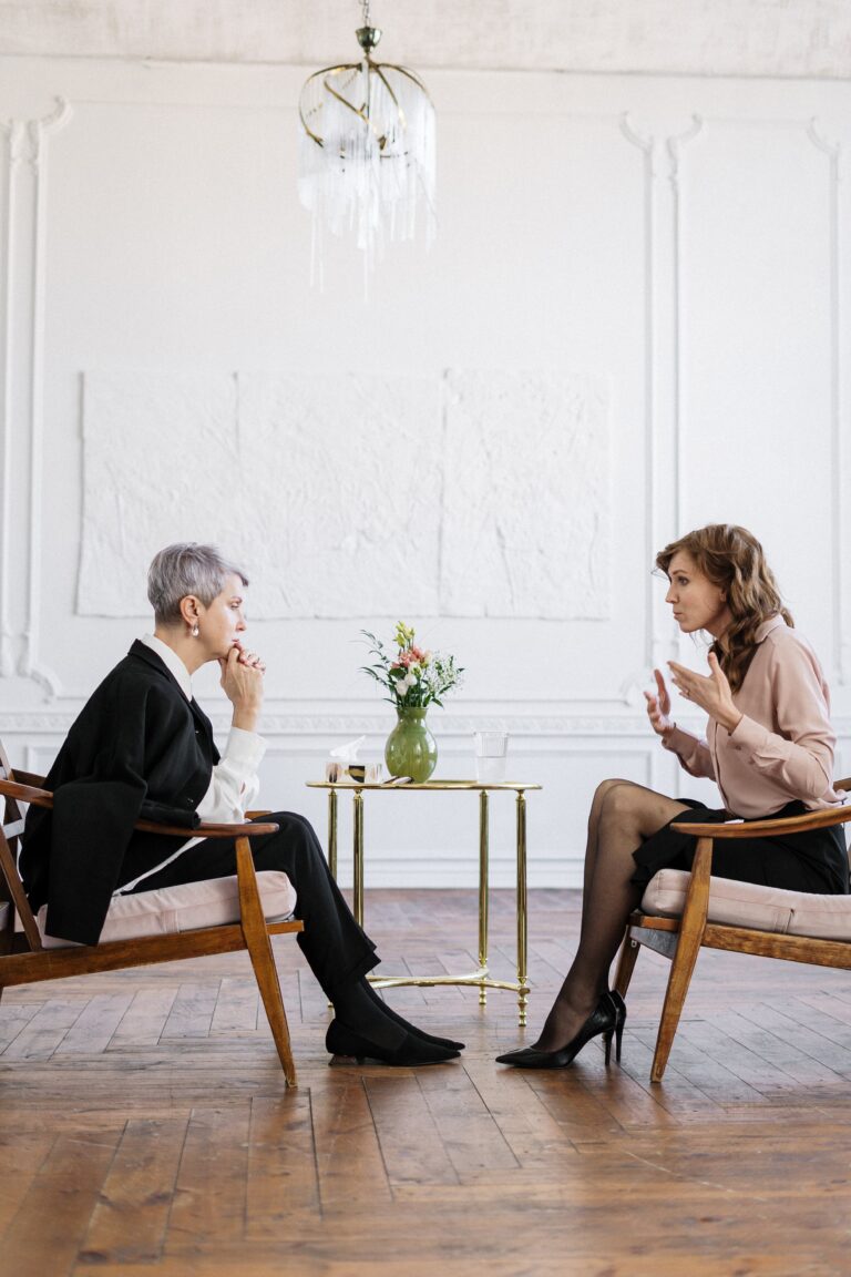Two women sit facing each other in wooden chairs, engaged in a serious conversation about the best Lead Magnet Tip. A small round table with a vase of flowers is between them in a bright, elegant room with wooden floors and white walls.