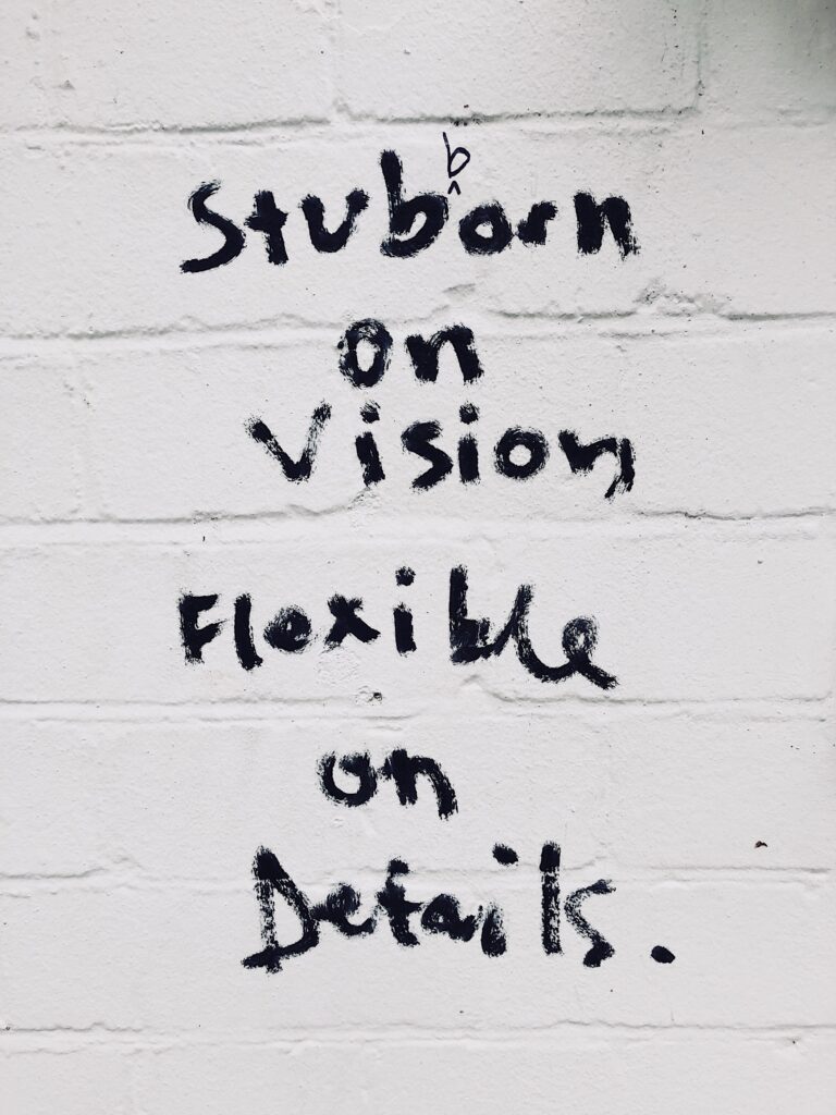 Black handwritten text on a white brick wall reads: Stubborn on vision, flexible on details. The writing appears uneven and informal, resembling graffiti—reminding you to let having a vision direct your steps.