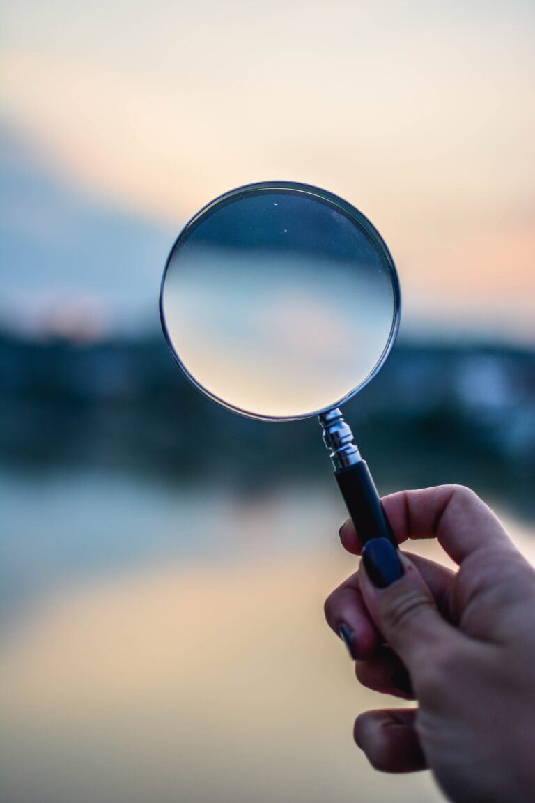 A hand with dark nail polish holds a magnifying glass outdoors, symbolizing customer clarity as it focuses on a blurred background with soft, pastel colors at sunset or dusk.