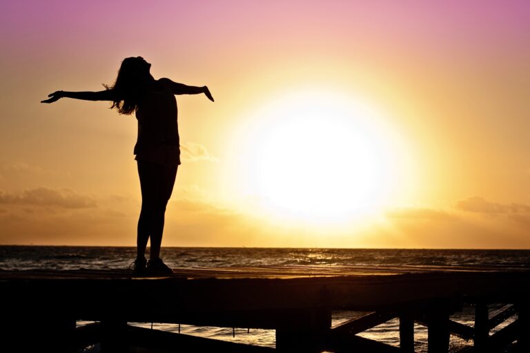 A person stands on a pier with arms outstretched, silhouetted against a vibrant sunset over the ocean, as if ready to break free beneath a sky glowing orange, yellow, and purple.