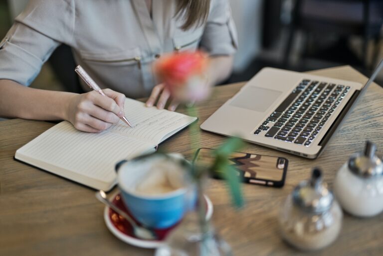 A person writes in a notebook next to an open laptop, a smartphone, and coffee on a wooden table, planning content marketing strategies in a cozy café setting to effectively reach audience goals.
