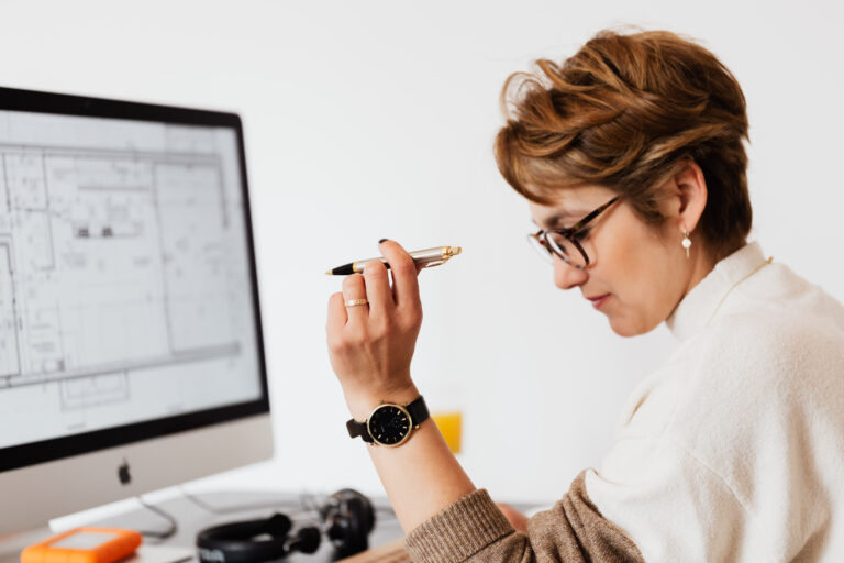 A decision-maker wearing glasses and a watch holds a pen, looking thoughtfully at architectural blueprints on a computer screen at their desk.