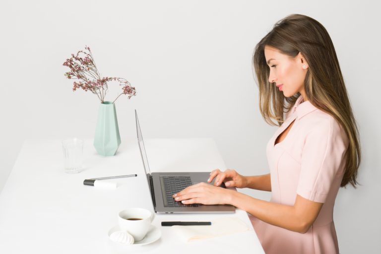 A woman in a light pink dress sits at a white desk working on strategic activities on her laptop. On the desk are a mint green vase with dried flowers, a cup of coffee, a pen, and a glass of water.